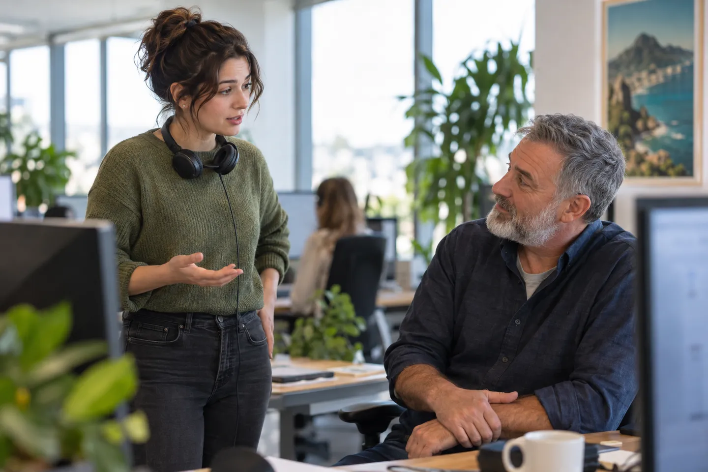 A support agent with a headset asks a question at a colleague's desk as he turns from his screen to listen.