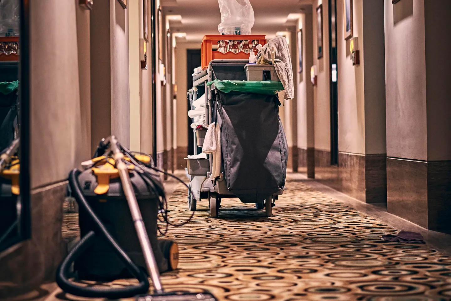A housekeeping trolley and a vacuum cleaner stand in a long, warmly lit hotel corridor with patterned carpet.