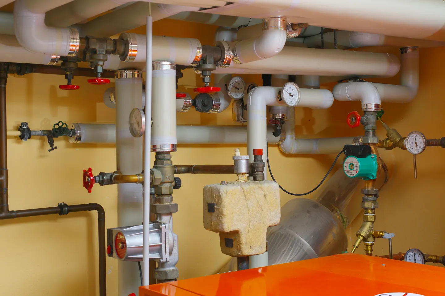 A boiler room with insulated pipes, red-handled valves and pressure gauges above an orange boiler and a green circulation pump.
