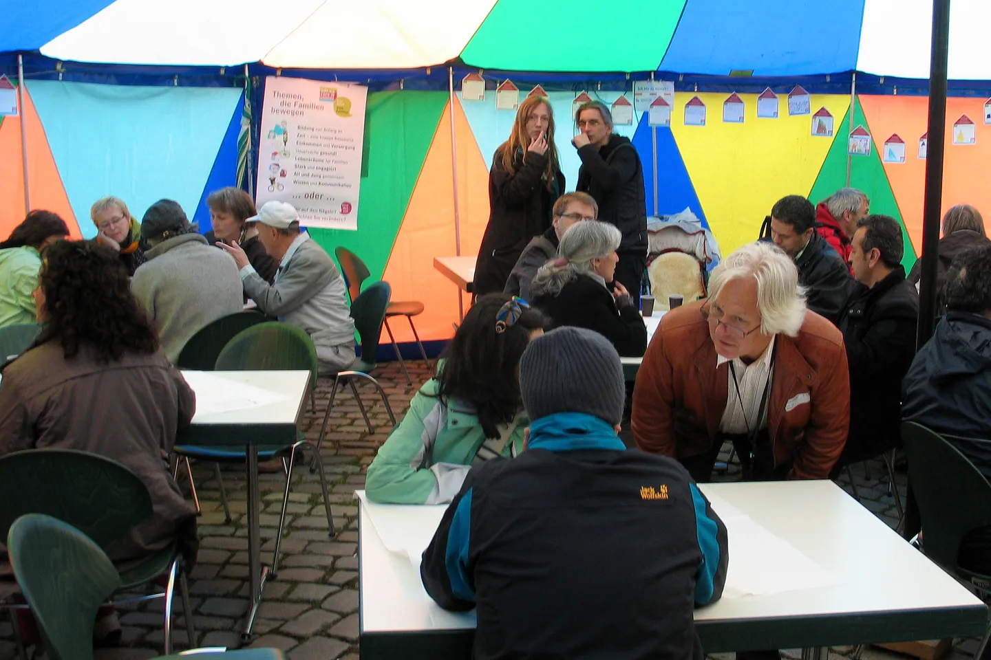 Small café tables in a striped festival tent, with groups of adults talking during a World Café.