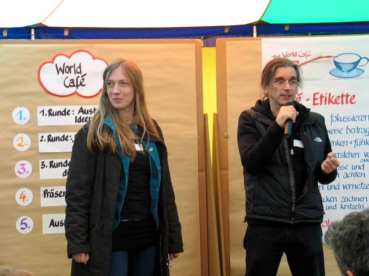 Two hosts, one speaking into a microphone, stand in a tent in front of boards listing numbered café rounds and a World Café etiquette poster.