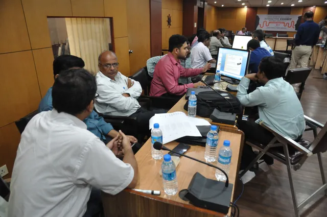 Workshop participants discuss ideas in groups at computer-equipped tables in a large room.