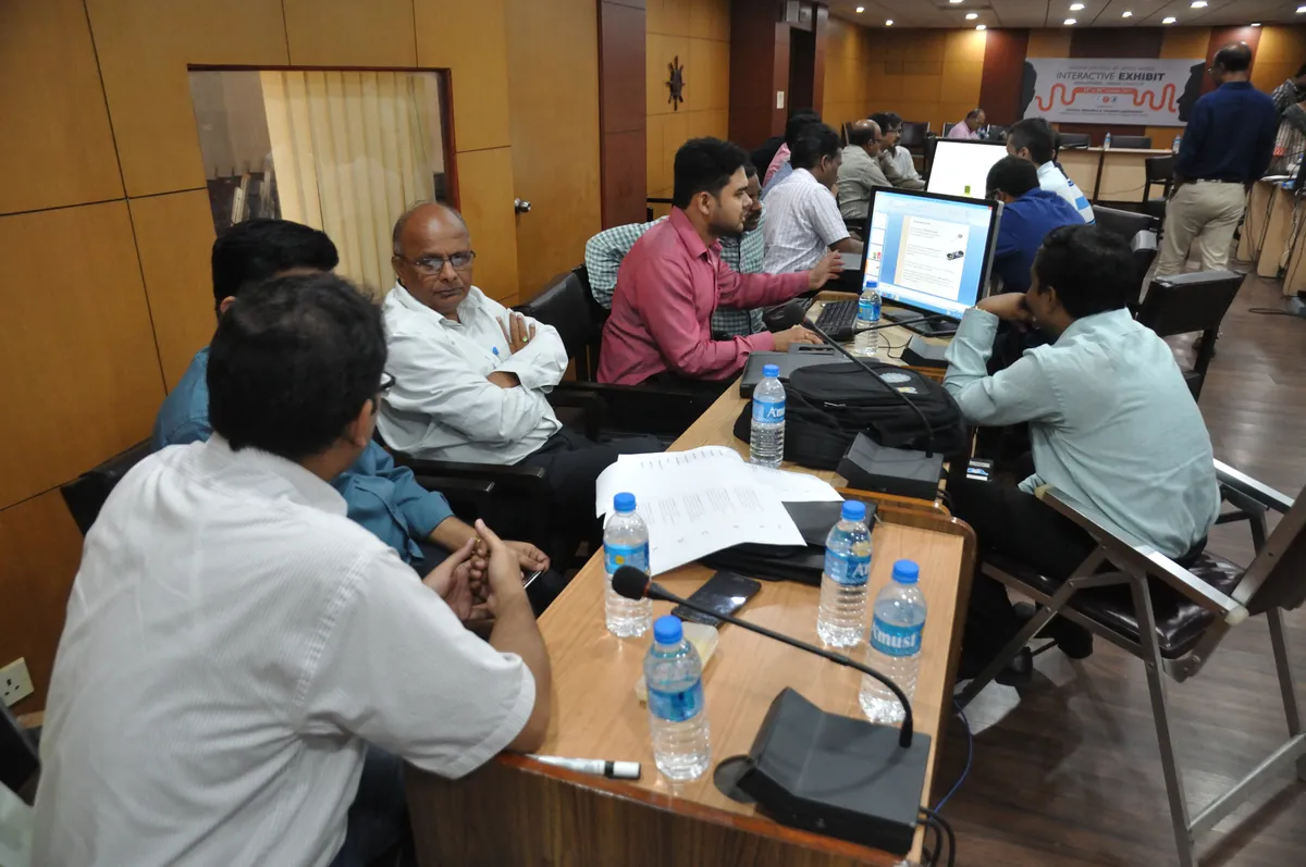 Workshop participants discuss ideas in groups at computer-equipped tables in a large room.