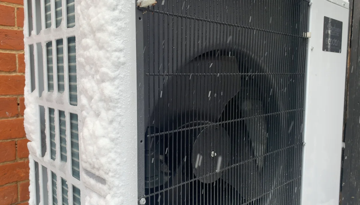 Close-up of an air-source heat pump's outdoor unit beside a brick wall, its side grille frosted with snow as flakes fall.