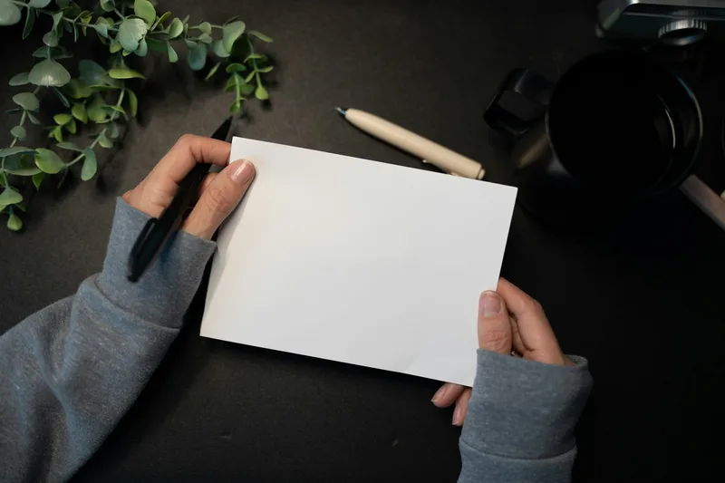 Hands holding a blank sheet of paper beside a pen, a mug and leafy stems.