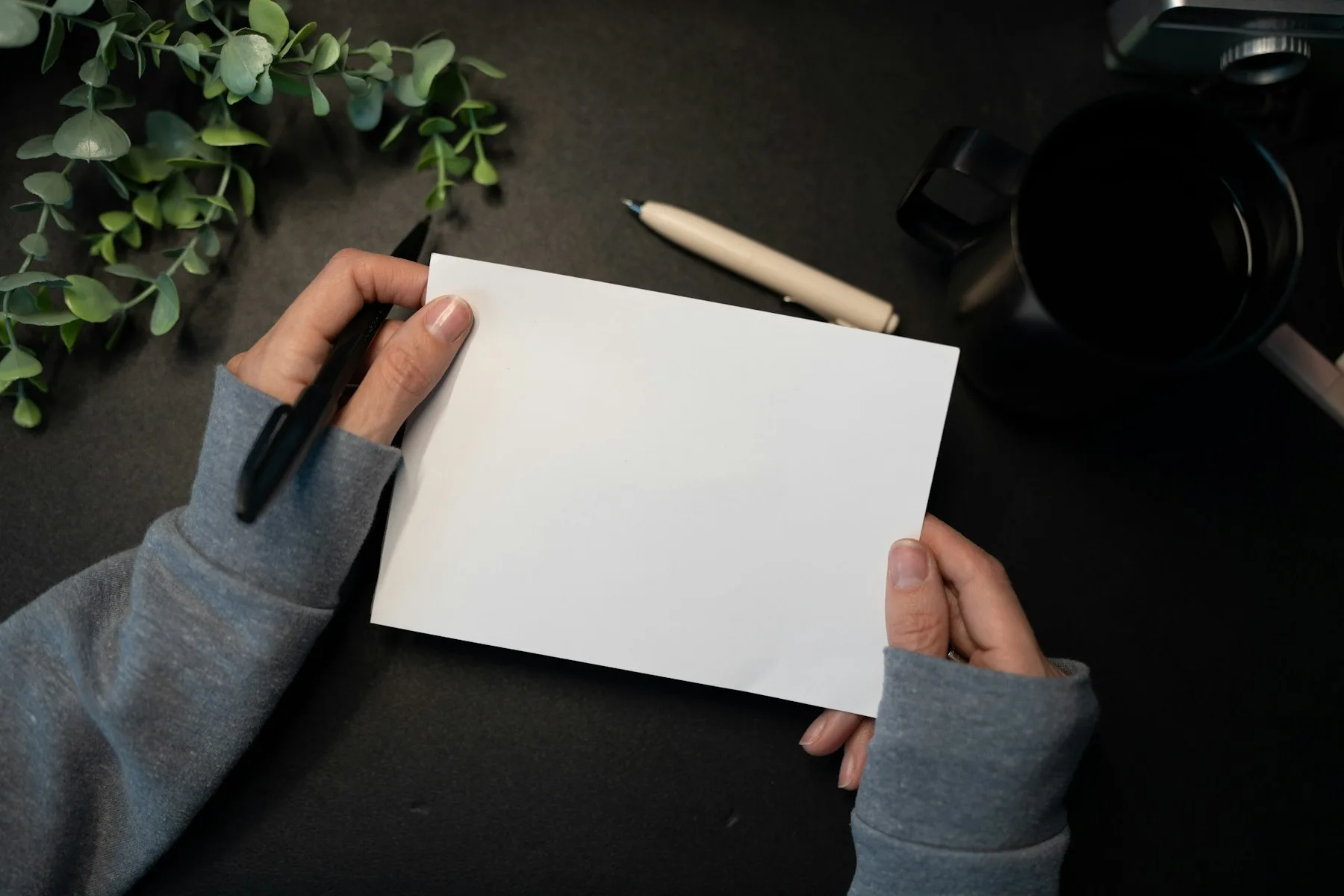 Hands holding a blank sheet of paper beside a pen, a mug and leafy stems.