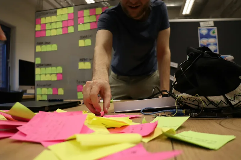 A person reaches across a working table for colourful sticky notes, with an affinity diagram in the background.